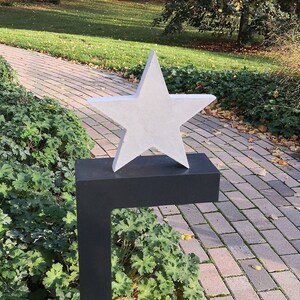 May include: A white, five-pointed star sculpture on a black metal stand. The star is positioned on a brick pathway in a garden setting.