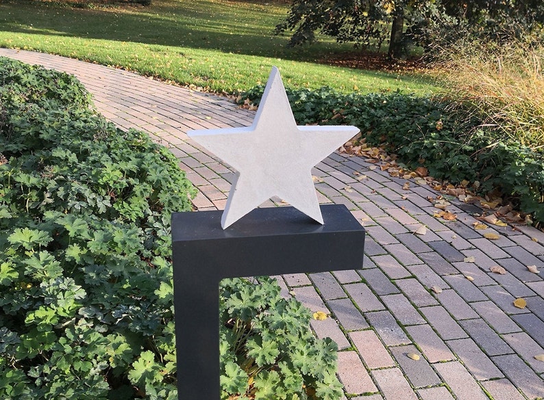 May include: A white, five-pointed star sculpture on a black metal stand. The star is positioned on a brick pathway in a garden setting.