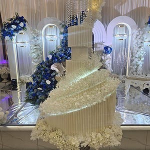 May include: A multi-tiered white cake with vertical fluted textures, featuring a spiraling floral arrangement of white blossoms, integrated LED lighting, and a crystal chandelier-style separator, all displayed on a silver wire-frame pedestal base.