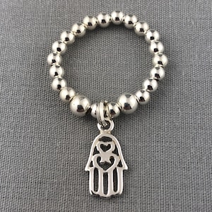May include: A silver beaded ring with a hamsa hand charm. The hamsa hand charm has a cutout design with three hearts in the centre.
