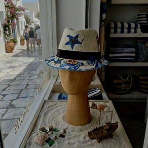 May include: A straw fedora-style hat with a black band and a blue and white sea life design. The hat is displayed on a wooden mannequin head in a shop window. The shop window also displays a small wooden boat and seashells.
