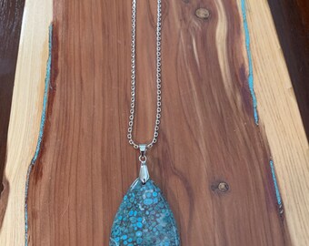 May include: A silver chain necklace with a large, teardrop-shaped pendant. The pendant features a mottled pattern of teal, turquoise, and white, resembling a gemstone. The necklace is displayed on a wooden surface.