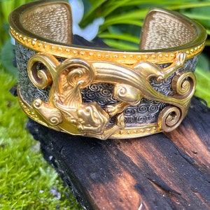 May include: A gold and black bracelet with a detailed design of a mythical creature. The bracelet is made of metal and has a textured surface.