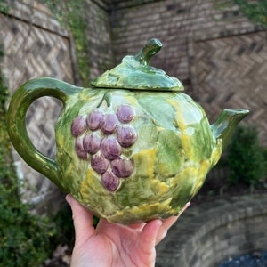 May include: A ceramic teapot with a green and yellow leaf design and a cluster of purple grapes. The teapot has a curved handle, a spout, and a lid with a stem-like handle. The teapot is held in a hand.