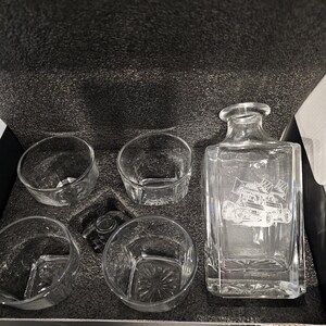 May include: A clear glass decanter with a square base and four glasses. The decanter has an etched design of a race car. The set is packaged in a black box with foam padding.
