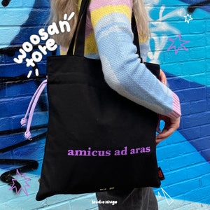 Peut inclure: Un sac fourre-tout noir avec des sangles violettes et le texte "amicus ad aras" imprimé en violet sur le devant. Le sac est porté par une personne vêtue d'un pull rayé pastel.