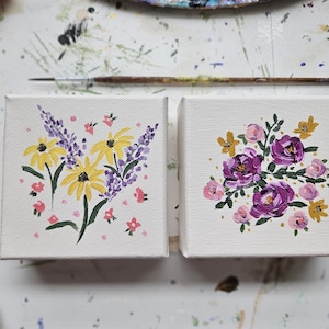 May include: Two square canvases featuring hand-painted floral designs. The left canvas displays yellow coneflowers and purple lavender sprigs with small pink accents. The right canvas shows a cluster of purple roses and small yellow flowers.