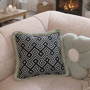 Peut inclure: Un coussin décoratif avec un motif géométrique noir et blanc et une bordure crochetée vert clair. Un coussin en forme de fleur de couleur vert doux est à côté. Les coussins sont sur un canapé beige.