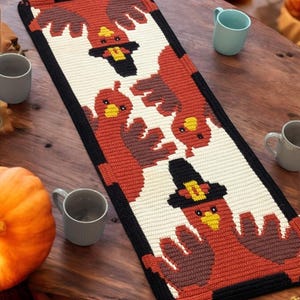 Peut inclure: Un chemin de table crocheté sur le thème de Thanksgiving. Le chemin de table présente un fond blanc avec une bordure noire et un motif répétitif de dindes avec des accents bruns, orange et jaunes.