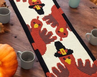 Puede incluir: Un camino de mesa tejido a crochet con temática de Acción de Gracias. El camino de mesa presenta un fondo blanco con un borde negro y un patrón repetitivo de pavos con acentos marrones, naranjas y amarillos.