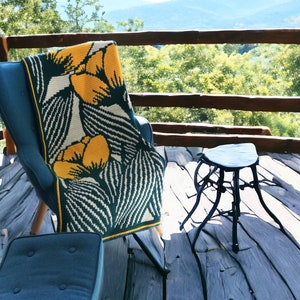 Op de afbeelding: Een groen-wit gecrochet deken met gele bloemenpatronen is over een blauwe stoel gegooid op een houten terras met uitzicht op een berg.