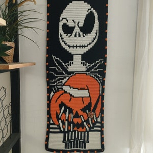 May include: A black and white crocheted wall hanging featuring Jack Skellington from The Nightmare Before Christmas holding a pumpkin. The hanging is framed with orange and black yarn.