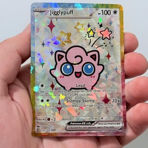 May include: A holographic Jigglypuff Pokémon trading card. The pink character has blue eyes and a smiling expression. The card features the text "Jigglypuff", "Lead", and "Stompy Stomp". The card has a holographic background with stars.