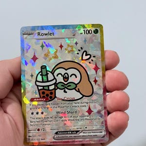 May include: A holographic Rowlet Pokémon card with a cartoon owl character holding a bubble tea. The card has a sparkly background with stars and hearts. The text on the card includes "Rowlet", "Sky Circus", and "Wind Shard".