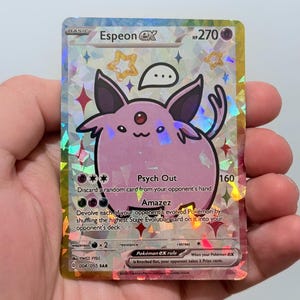 May include: A holographic Espeon ex Pokémon trading card. The card features a pink Espeon character with black ears and a red gem. The card has the text "Espeon ex" and "Psych Out" and "Amazez".
