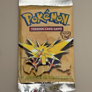 Può includere: Confezione sigillata di carte collezionabili Pokémon con un'illustrazione di Zapdos giallo. La confezione ha uno sfondo marrone con il logo Pokémon, il testo "Trading Card Game" e "Fossil". C'è scritto "11 Additional Game Cards".