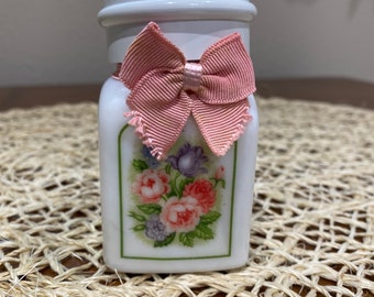 Puede incluir: Un tarro de cerámica blanco con un diseño floral y un lazo rosa en la tapa. El tarro está sobre un mantel tejido.