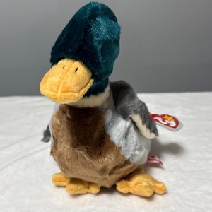 May include: A plush duck toy with a teal head, yellow beak, and brown and gray body. The duck has a red tag attached to its side. The toy is designed to resemble a mallard duck.