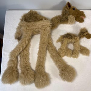 May include: Two plush camel toys with long, shaggy tan and brown fur. The larger camel features a prominent hump, a long neck, and dark eyes, while the smaller camel is a miniature version with similar shaggy fur, a small hump, and dark eyes.
