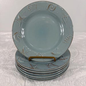 May include: A stack of six light blue ceramic plates featuring embossed seashell and starfish designs along the rim. Each plate has a smooth center and a textured border with brown detailing highlighting the raised marine motifs.