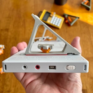 Peut inclure: Un lecteur de musique portable blanc avec un support gris, une ligne rouge et un bouton. Le lecteur possède une prise casque, une prise microphone, un port USB et un bouton sur le côté.