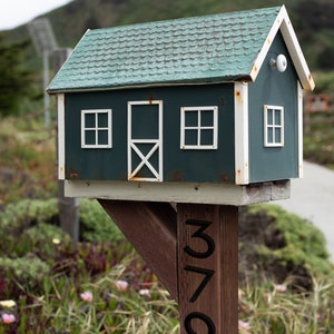 May include: A green wooden mailbox shaped like a house with white trim and a white door. The mailbox is mounted on a wooden post with the number 3795 painted on it.