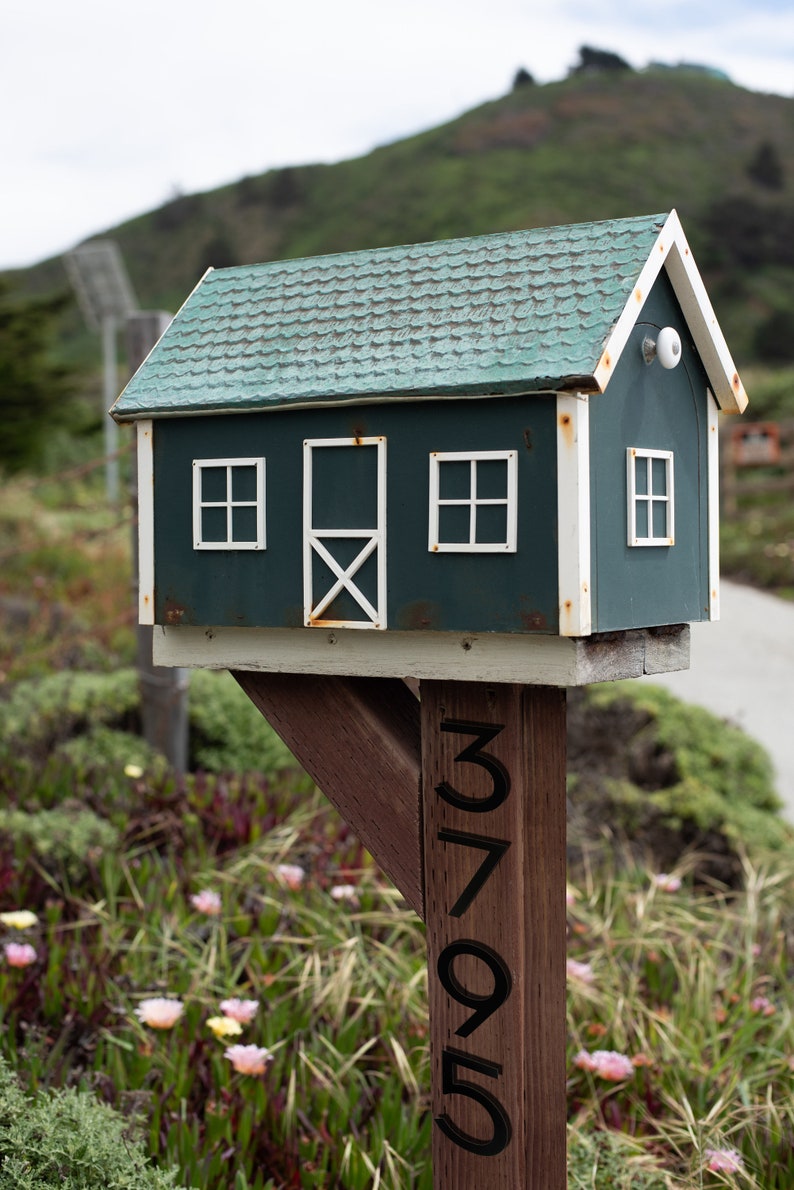 May include: A green wooden mailbox shaped like a house with white trim and a white door. The mailbox is mounted on a wooden post with the number 3795 painted on it.
