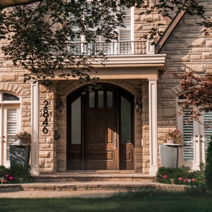 May include: A brown wooden front door with a glass panel and a black door handle. The door is framed by a stone archway and is surrounded by stone walls. The house number 2846 is displayed on the wall to the left of the door.