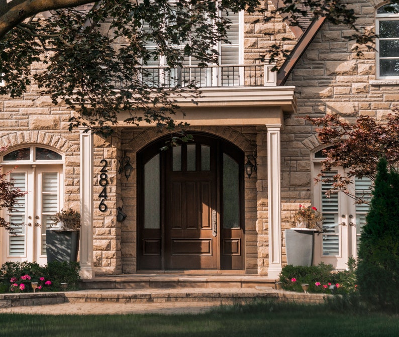 May include: A brown wooden front door with a glass panel and a black door handle. The door is framed by a stone archway and is surrounded by stone walls. The house number 2846 is displayed on the wall to the left of the door.
