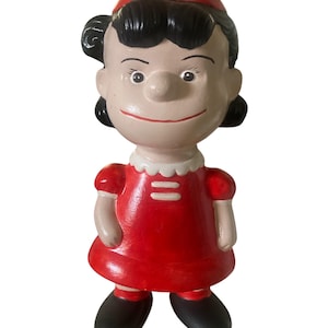 May include: A ceramic figurine of a cartoon girl with black hair, a red hat, and a red dress. The figurine has a smiling expression and black shoes. The dress has a white collar with two horizontal white stripes.