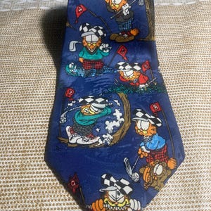 May include: A blue necktie featuring a repeating pattern of Garfield playing golf. The cartoon cat is depicted in various golf poses, wearing a checkered hat and golf attire. The tie has a pointed end.