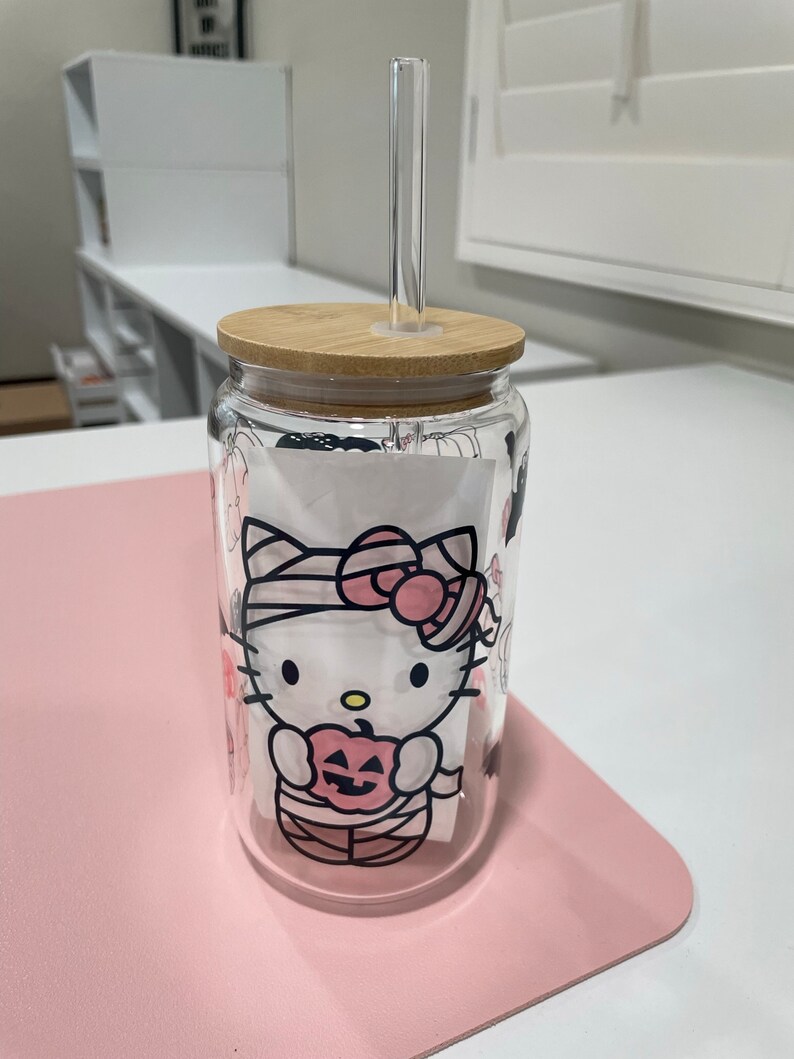 May include: A clear glass tumbler with a bamboo lid and a Hello Kitty Halloween design. The design features Hello Kitty dressed as a mummy holding a jack-o-lantern. The tumbler has a clear plastic straw.