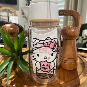 May include: A clear glass water bottle with a bamboo lid and a straw. The bottle has a Halloween-themed design featuring Hello Kitty dressed as a mummy holding a pumpkin.