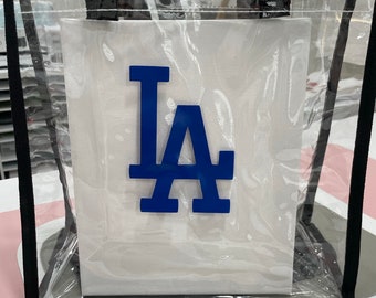 May include: Clear plastic tote bag with black handles and a white canvas insert featuring a blue "LA" logo.