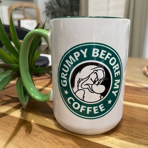 May include: White ceramic coffee mug with a green rim and a green circle design. The circle features a cartoon character with a grumpy expression and the text "Grumpy Before My Coffee".