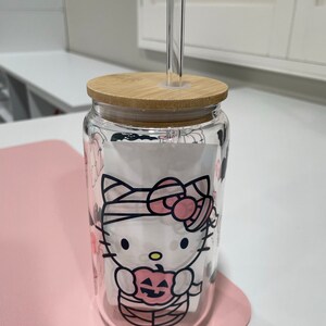 May include: A clear glass tumbler with a bamboo lid and a Hello Kitty Halloween design. The tumbler has a clear glass straw and is sitting on a pink surface.