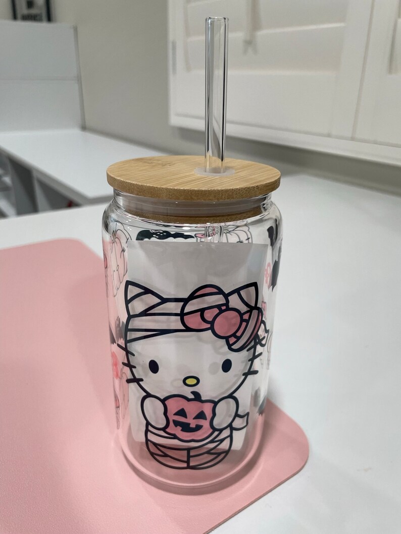 May include: A clear glass tumbler with a bamboo lid and a Hello Kitty Halloween design. The tumbler has a clear glass straw and is sitting on a pink surface.