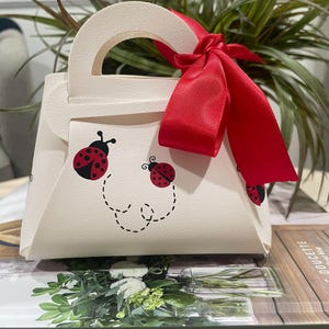 May include: A cream-colored gift bag with a handle and a red satin ribbon tied in a bow. The bag is decorated with ladybug graphics and a dotted line design. The bag is sitting on a book with the text "GIFT OF HOME".