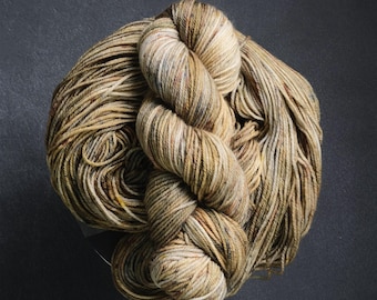 May include: A single skein of hand-dyed yarn featuring a variegated color palette of earthy tan, muted olive green, and soft brown tones. The twisted fiber displays subtle speckles of rust and cream throughout the tightly spun strands.