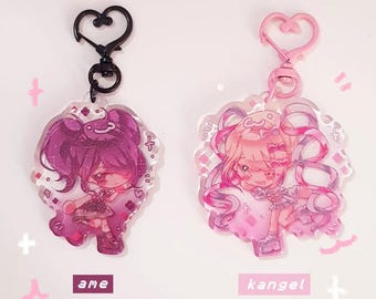 May include: Two acrylic charms featuring anime-style characters. One charm has a black heart-shaped clasp, the other a pink heart-shaped clasp. The charms are approximately 2.5 inches with glitter epoxy. The text "ame" and "kangel" are visible.