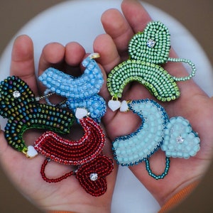 May include: Five beaded, colorful, decorative ornaments in the shape of a stylized butterfly. Each ornament has a small, clear rhinestone in the center. The ornaments are held in a person's hand.