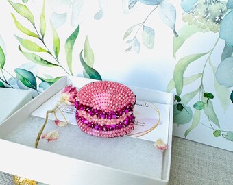 Puede incluir: Un amuleto de macaron de cuentas rosa y morado en una caja de regalo blanca. La caja está sobre una superficie de mármol con detalles dorados.