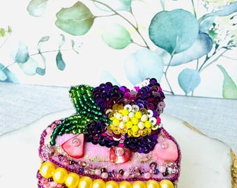 Puede incluir: Un macaron de cuentas rosa y morado con una flor amarilla y blanca en la parte superior. El macaron está decorado con cuentas rosas, blancas y amarillas. El macaron está sobre una superficie blanca con detalles dorados.