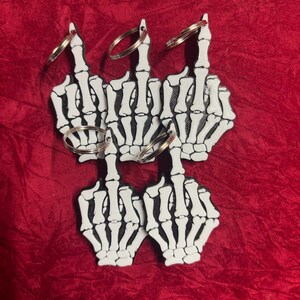 Könnte beinhalten: Fünf Schlüsselanhänger in Form von Skeletthänden, die den Mittelfinger zeigen. Die Schlüsselanhänger sind weiß mit schwarzen Details und haben silberne Schlüsselringe. Der Hintergrund ist einfarbig rot.