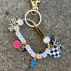 May include: A gold-toned keychain with a variety of charms. Charms include a checkered flag, a pink smiley face, a motorcycle, and a blue lightning bolt. The word "MOTO" is spelled out in white letter beads.
