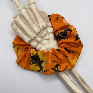 K&ouml;nnte beinhalten: Ein orangefarbener und schwarzer Stoff-Haargummi mit einem Hexen- und Mondmuster. Das Haargummi ist um eine wei&szlig;e Skeletthand gewickelt.