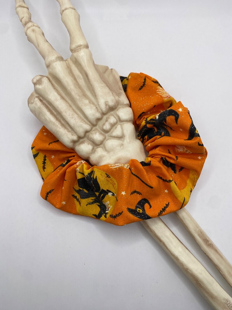 K&ouml;nnte beinhalten: Ein orangefarbener und schwarzer Stoff-Haargummi mit einem Hexen- und Mondmuster. Das Haargummi ist um eine wei&szlig;e Skeletthand gewickelt.