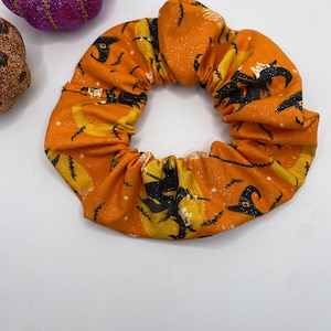 K&ouml;nnte beinhalten: Ein orangefarbener Stoff-Haargummi mit einem Halloween-Druck von Hexen, die auf Besen fliegen, Flederm&auml;usen und Sternen.
