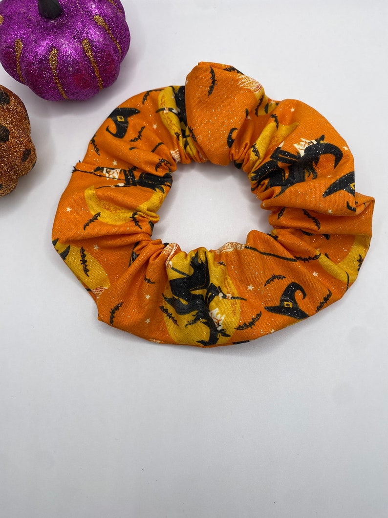 K&ouml;nnte beinhalten: Ein orangefarbener Stoff-Haargummi mit einem Halloween-Druck von Hexen, die auf Besen fliegen, Flederm&auml;usen und Sternen.