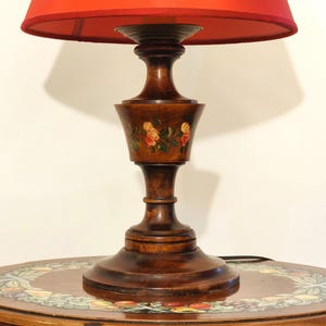 May include: A vintage wooden table lamp with a vibrant red lampshade. The lamp features a dark brown, ornate base with hand-painted floral details. The lamp is placed on a round wooden table with a decorative floral design.